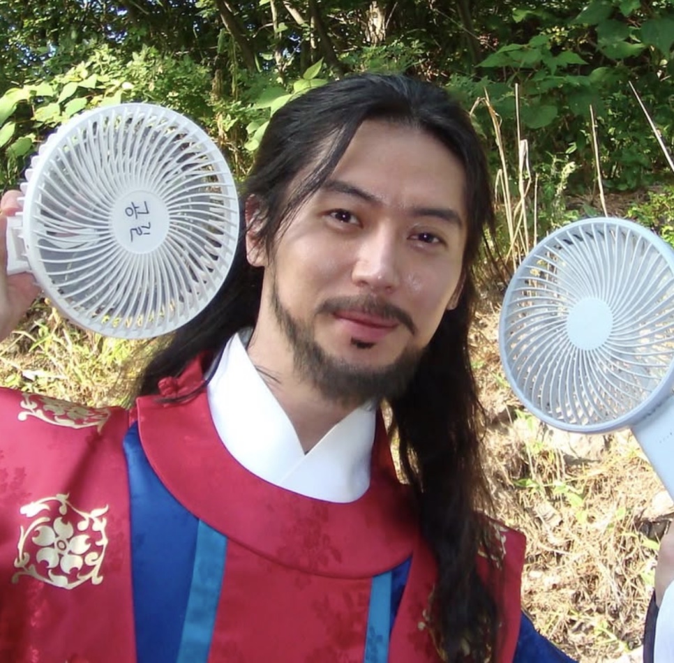Lee Joo-ahn de Gong-gil, com roupa tradicional vermelha, barba e cabelo comprido, entre dois ventiladores portáteis