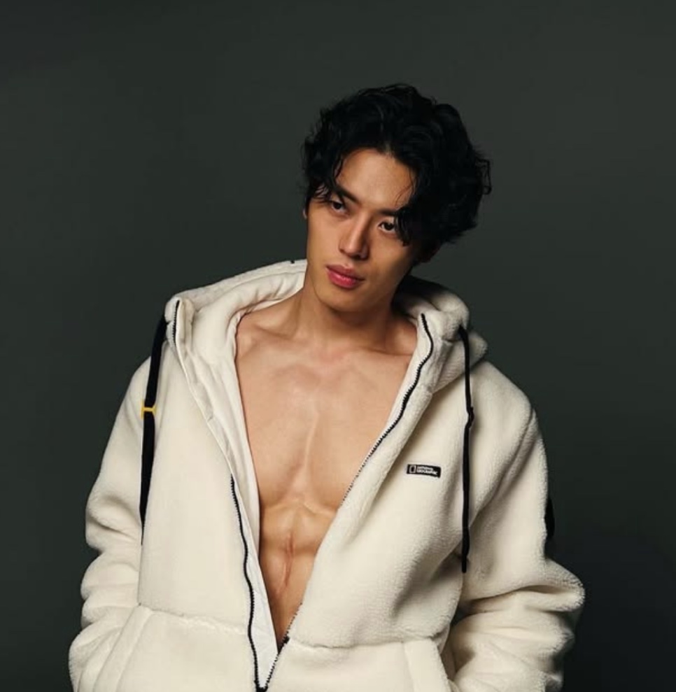 Lee Joo-ahn de casaco de pelúcia branco aberto, sem camisa, na frente de um fundo escuro
