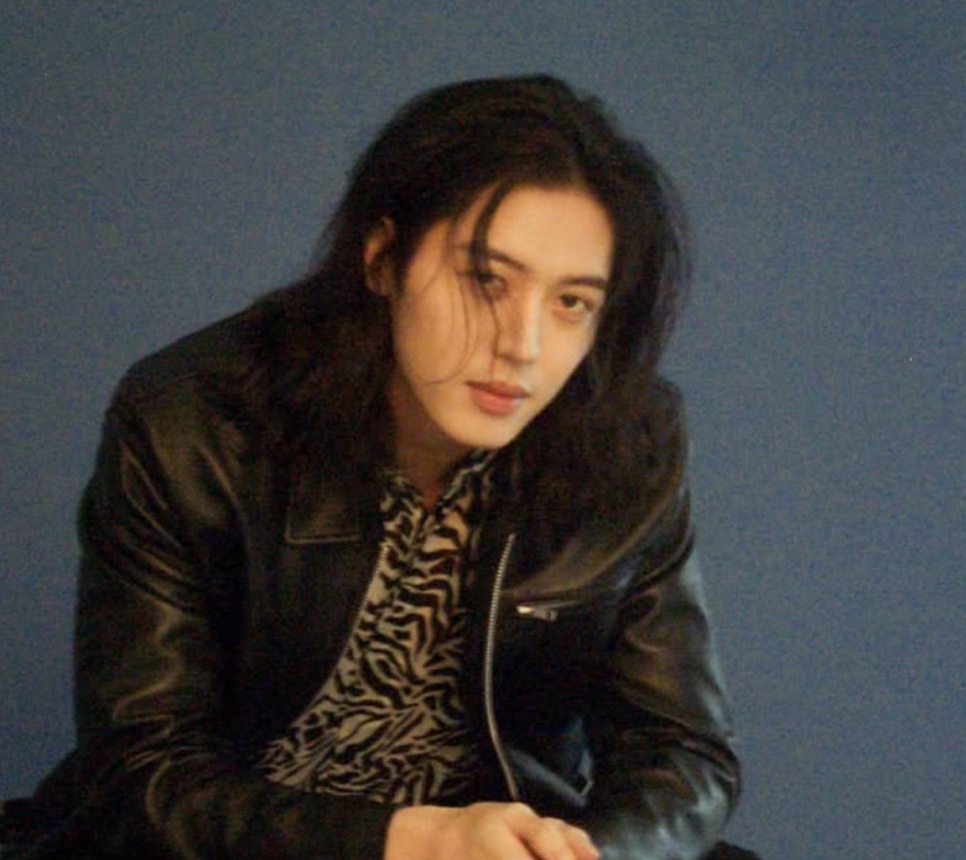 Lee Joo-ahn de cabelo comprido caindo no rosto, com jaqueta de couro preta sobre camisa de estampa animal