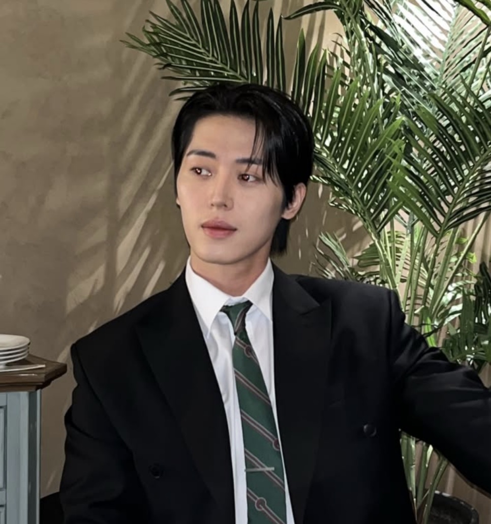 Lee Joo-ahn de terno preto e gravata verde listrada, olhando pro lado, na frente de uma palmeira