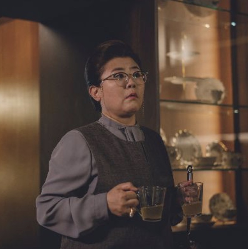 Lee Jung-eun como a governanta Moon-gwang, de óculos e colete cinza, segurando duas xícaras de chá em Parasita