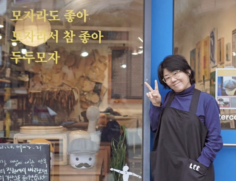 Lee Jung-eun de avental, fazendo sinal de paz na porta de uma loja de chapéus