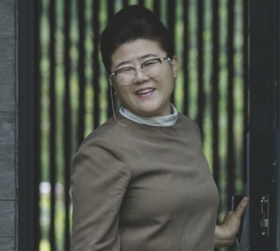 Lee Jung-eun como a governanta Moon-gwang, sorrindo na porta da casa em Parasita
