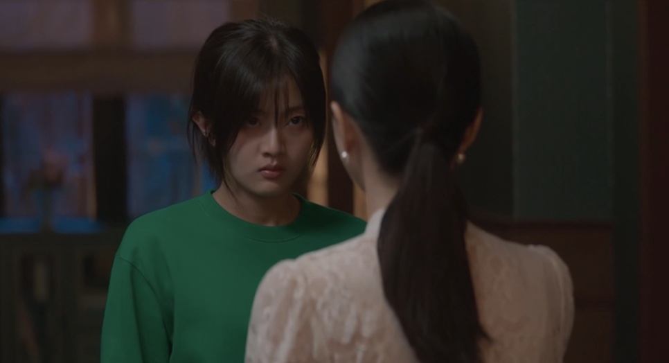 Yoon Cheong-a de camiseta verde, com cara fechada, atrás de uma mulher de cabelo preso