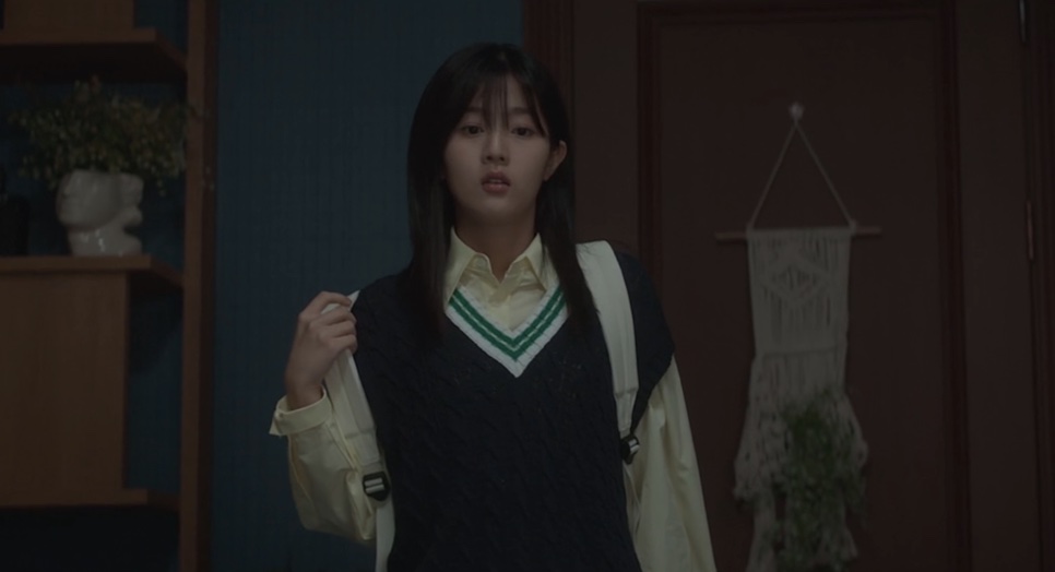 Yoon Cheong-a de colete escuro, segurando a alça da mochila, num quarto