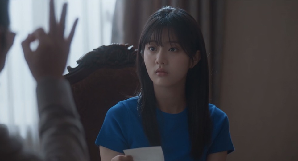 Yoon Cheong-a de blusa azul segurando um papel, enquanto alguém faz um sinal com a mão
