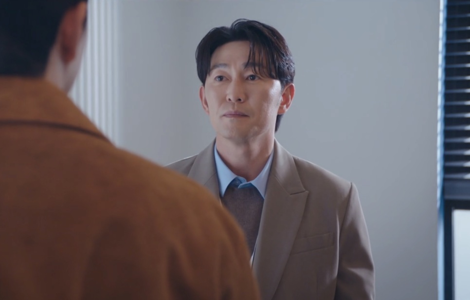 Choi Seong-uk de blazer bege, conversando com alguém num escritório