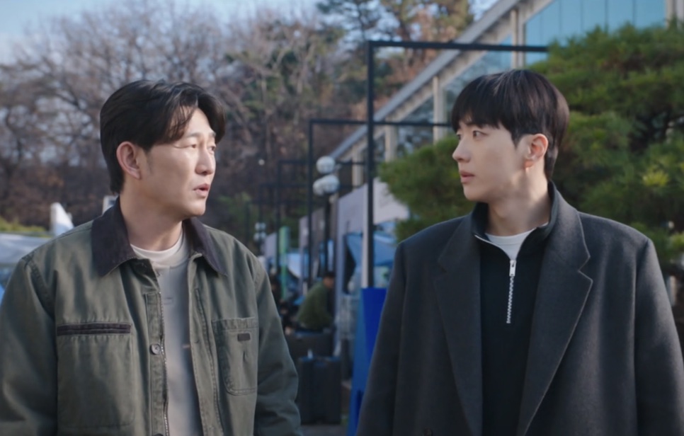 Choi Seong-uk de jaqueta verde ao lado do Kang Ha-gi, de casaco cinza, na frente de um prédio