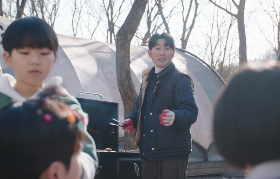 Choi Seong-uk de colete acolchoado, fazendo churrasco ao ar livre com crianças em volta