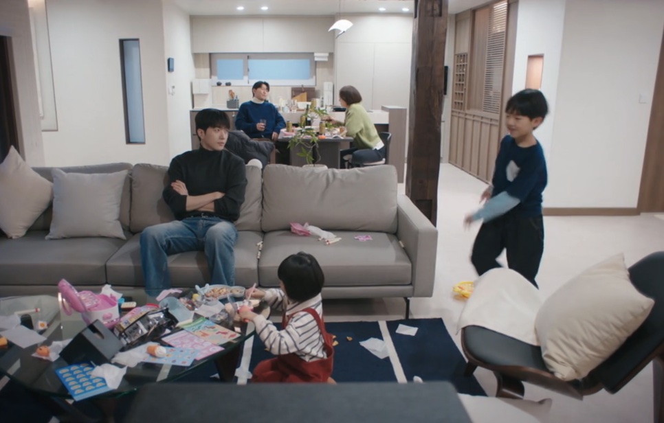 Kang Ha-gi sentado de braços cruzados num sofá, numa sala cheia de crianças brincando