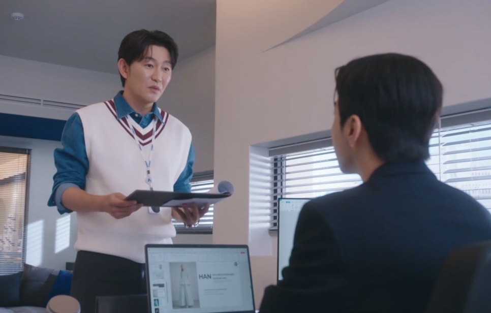 Choi Seong-uk em pé com uma pasta na mão, falando com o Kang Ha-gi sentado à mesa