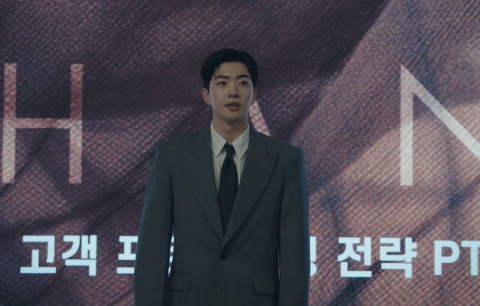 Kang Ha-gi de terno, em pé na frente de um telão durante uma apresentação