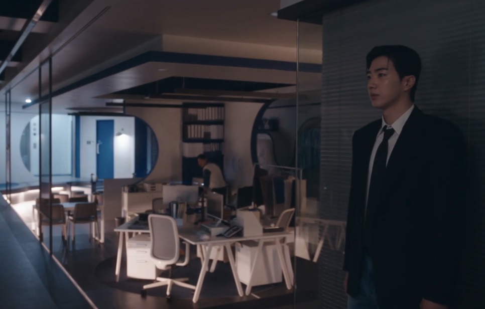 Kang Ha-gi de terno escuro, em pé na porta de um escritório vazio e com pouca luz