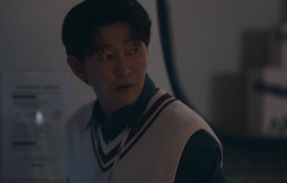Choi Seong-uk na penumbra, de colete branco com listras vinho, com o olhar abatido