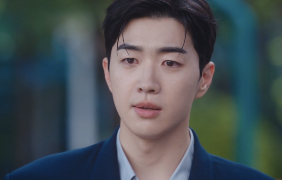 Close do Kang Ha-gi de terno azul-marinho, ao ar livre, com expressão triste