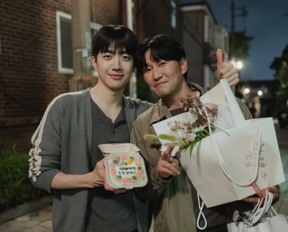 Kang Hoon e Heo Joon-seok sorrindo à noite, com um bolo e um buquê de flores