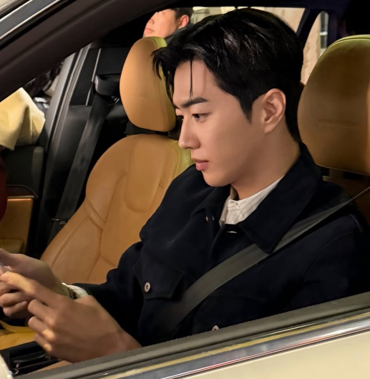Kang Ha-gi dentro de um carro, de casaco escuro, olhando o celular