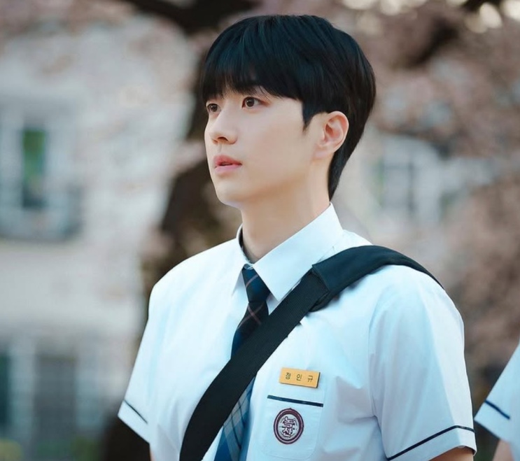 Kang Hoon de uniforme escolar, de perfil, com cerejeiras floridas ao fundo