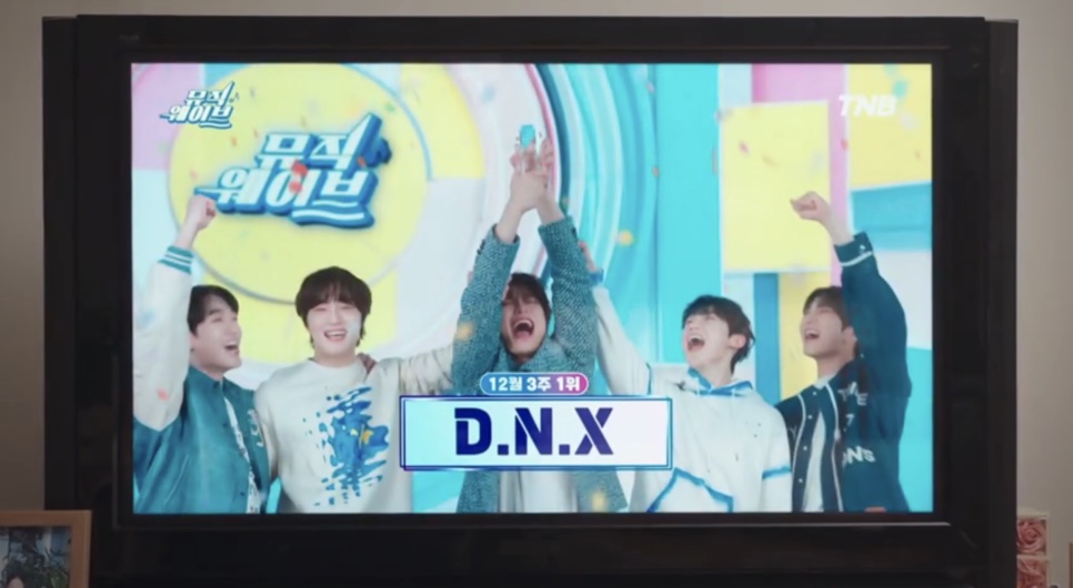 Uma TV mostrando os cinco integrantes do D.N.X comemorando o primeiro lugar num programa musical
