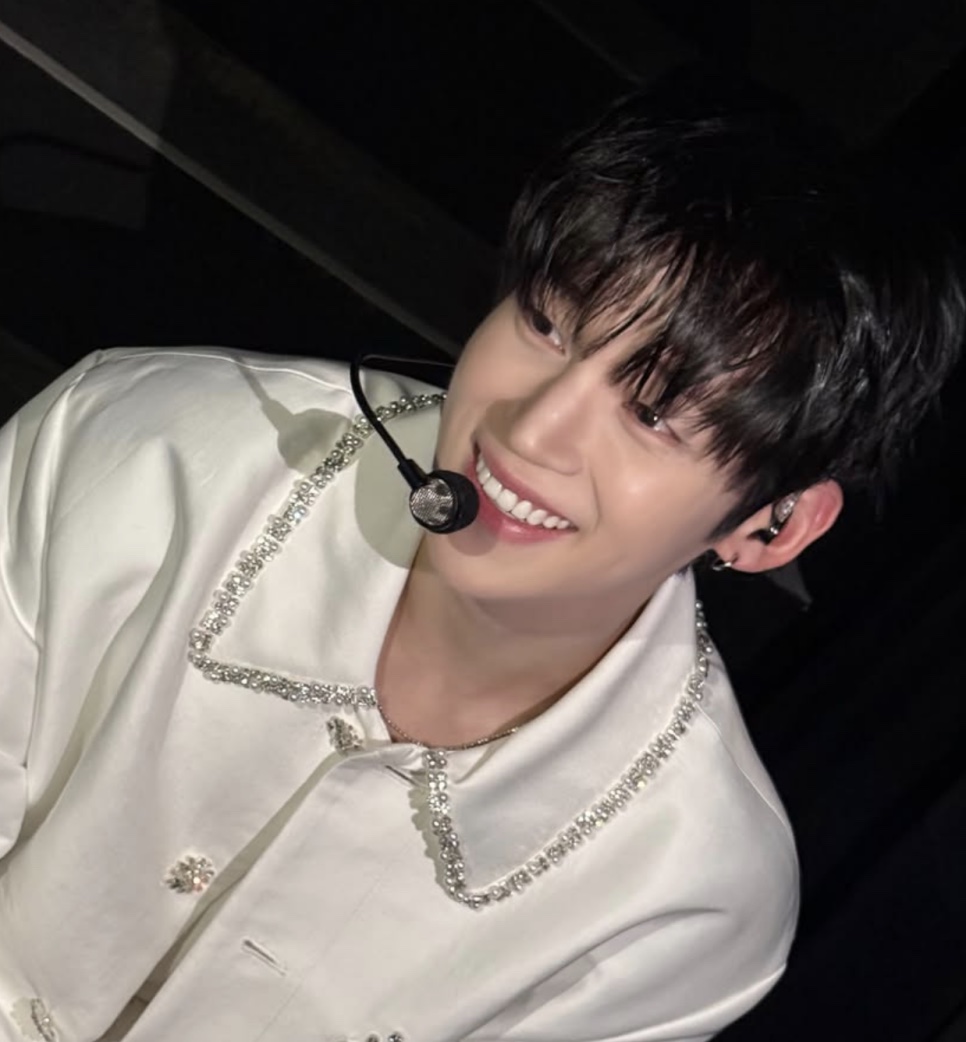 Um idol de casaco branco bordado, com microfone headset, sorrindo no palco