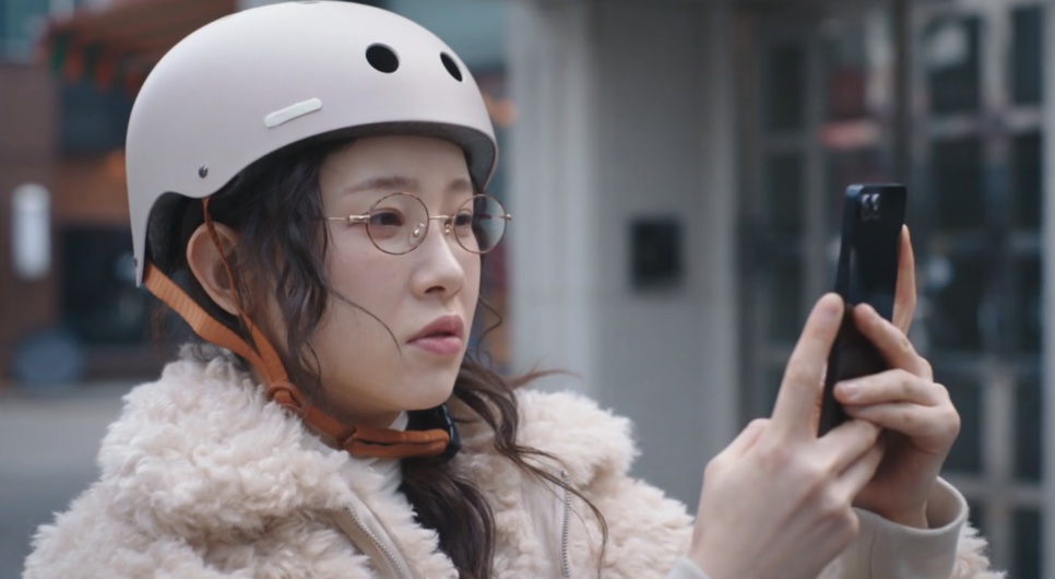 Uma mulher de capacete branco e óculos, na rua, olhando para o celular na mão