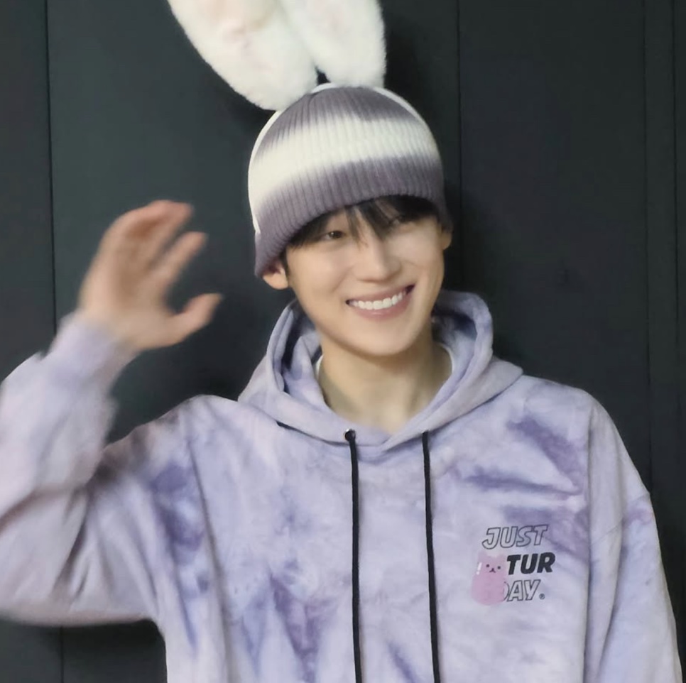 Um rapaz de gorro bege e moletom lilás sorrindo e acenando para a câmera