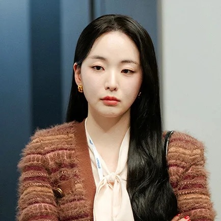 Seo Hye-su de cardigã felpudo cor de vinho e blusa com laço, num corredor de escritório