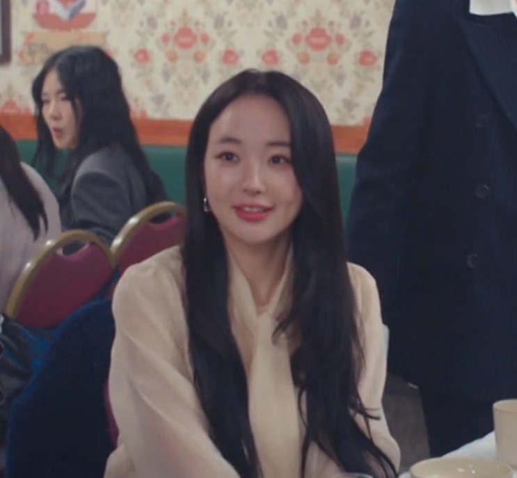 Seo Hye-su sorrindo, sentada a uma mesa de festa com papel de parede floral atrás