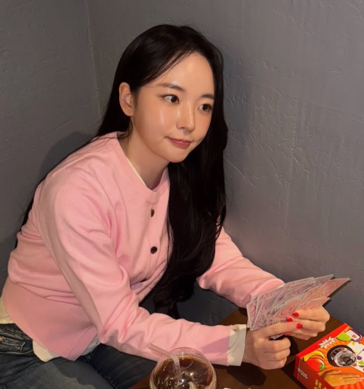 Kim Chae-eun de moletom rosa, sentada num canto de café segurando um envelope