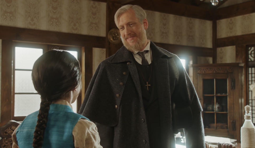 Joseph Stenson de cabelo grisalho e cruz no peito, conversando com uma pessoa de trança vista de costas