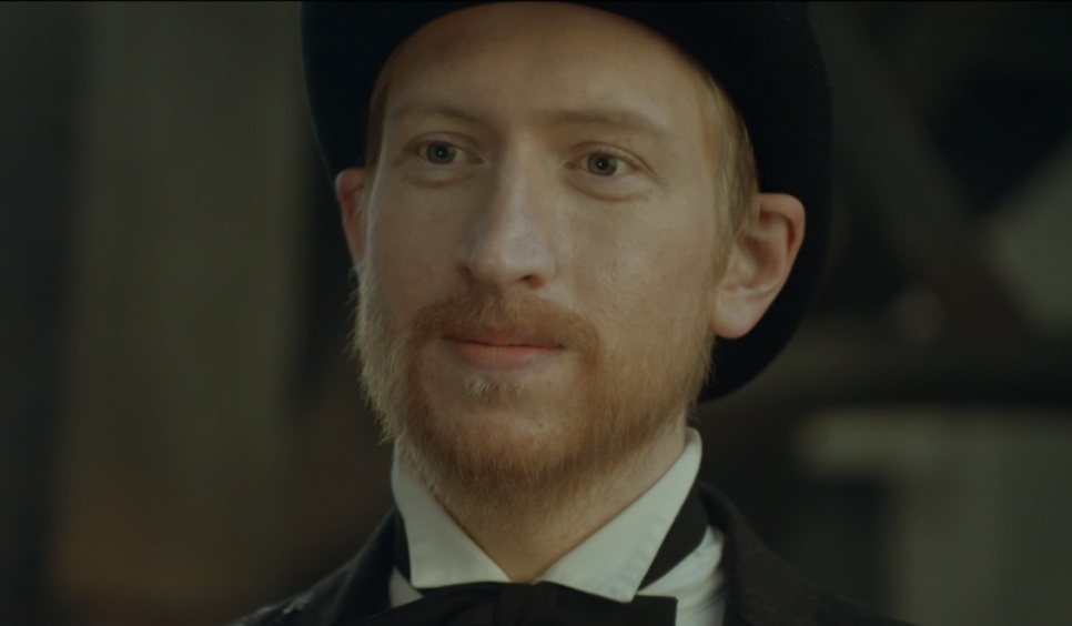 Close de Joseph Stenson de cartola, com barba ruiva, em um lugar escuro