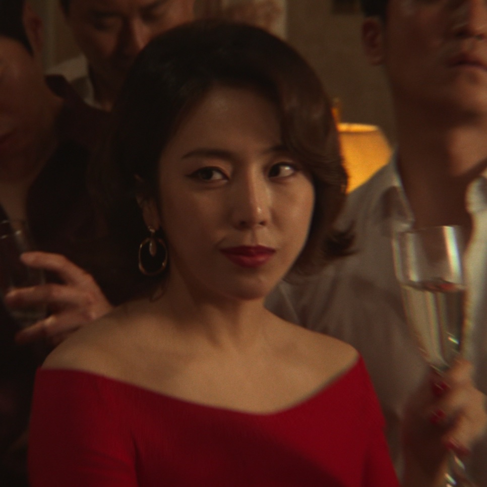 Yewon de vestido vermelho ombro a ombro e brincos de argola, com uma taça na mão, no papel da samonim de Narco-Santos