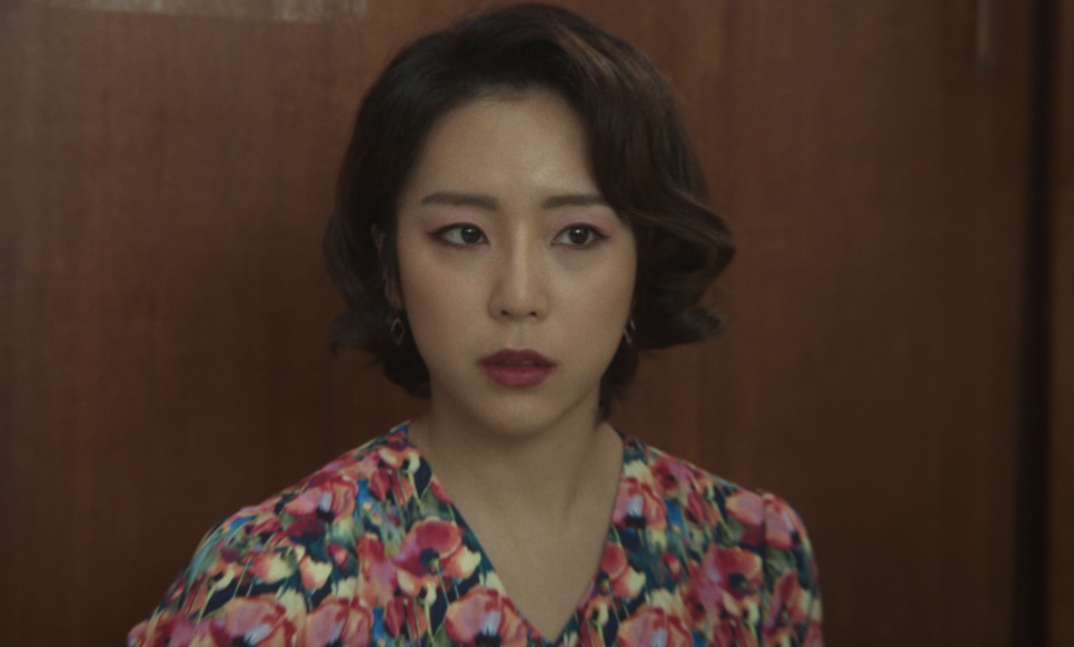 Yewon de vestido florido e cabelo curto ondulado, olhando para a frente com expressão séria, em Narco-Santos