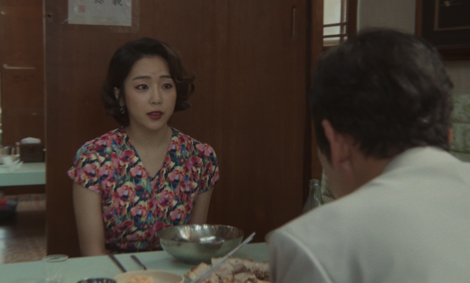 Yewon de vestido florido sentada à mesa de um restaurante, de frente para um homem de camisa branca, em Narco-Santos