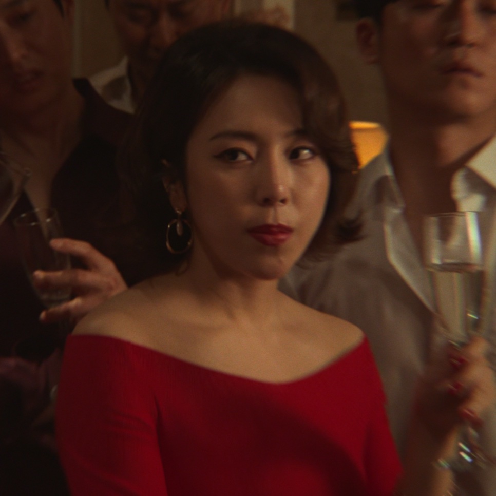 Close da Yewon de vestido vermelho e brincos de argola, no papel da samonim de Narco-Santos