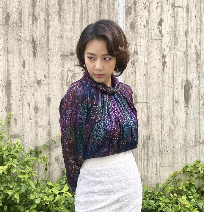 Yewon posando com o figurino da samonim, blusa brilhante estampada e saia branca, em frente a uma parede de concreto