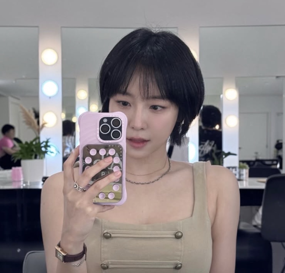 Yewon de cabelo curto tirando selfie no espelho de um camarim