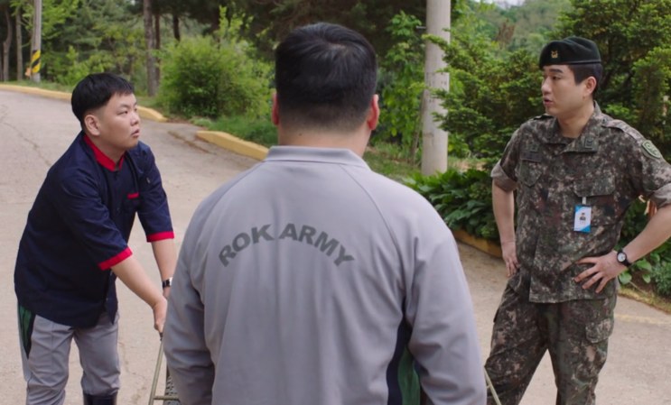 Kwaktube conversando com dois militares em uma via do quartel, um deles de costas com o agasalho do Exército coreano