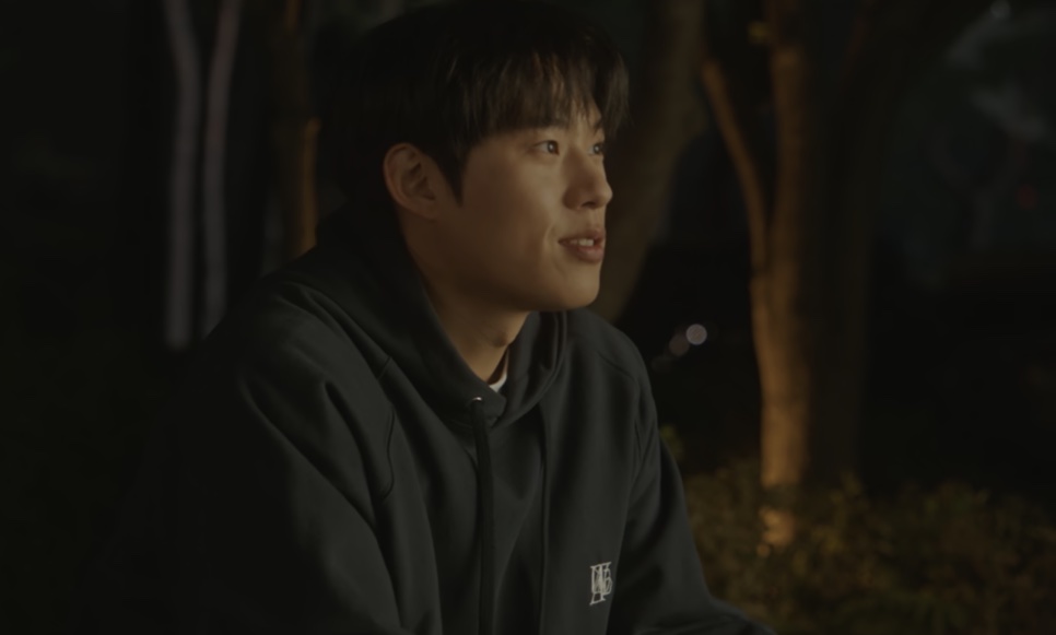 O Ji-ung de moletom preto, sentado ao ar livre à noite, sorrindo