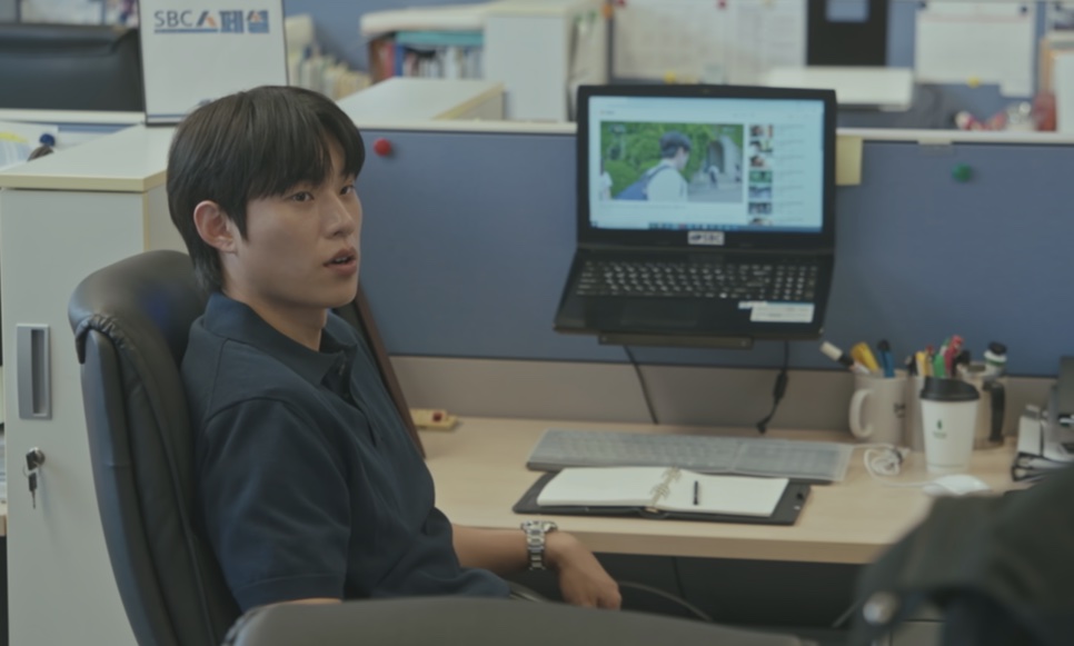 O Ji-ung sentado à mesa de trabalho, com um notebook aberto, no escritório da emissora
