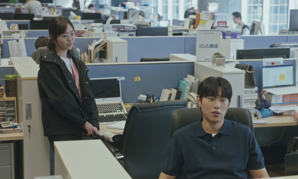 O Ji-ung sentado no escritório da emissora, com uma colega em pé ao lado da mesa
