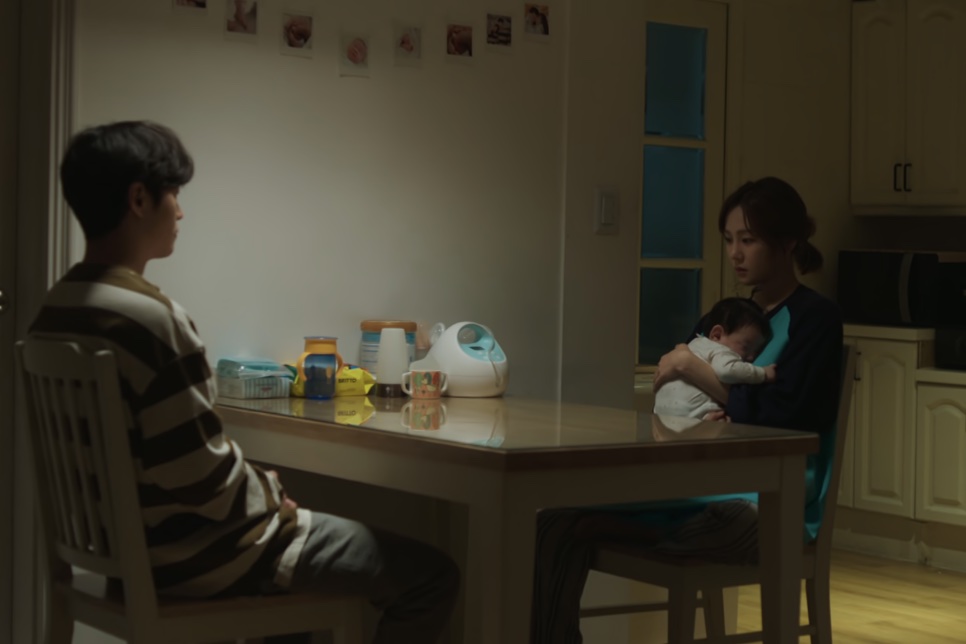 A Han-ju segurando o bebê à mesa da cozinha, com um homem sentado de costas do outro lado