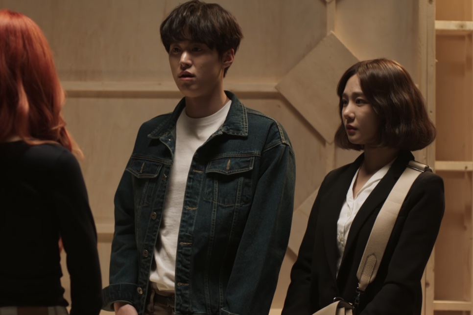 A Han-ju ao lado do Jae-hun, de jaqueta jeans, numa sala com paredes de madeira