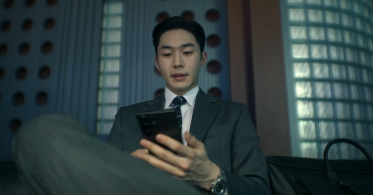 Son Soo-hyun sentado, olhando o celular em uma sala clara