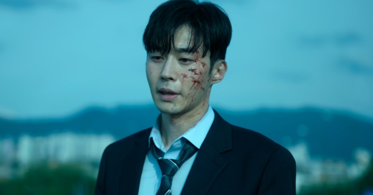 Son Soo-hyun com o rosto machucado, ao ar livre, com a cidade ao fundo