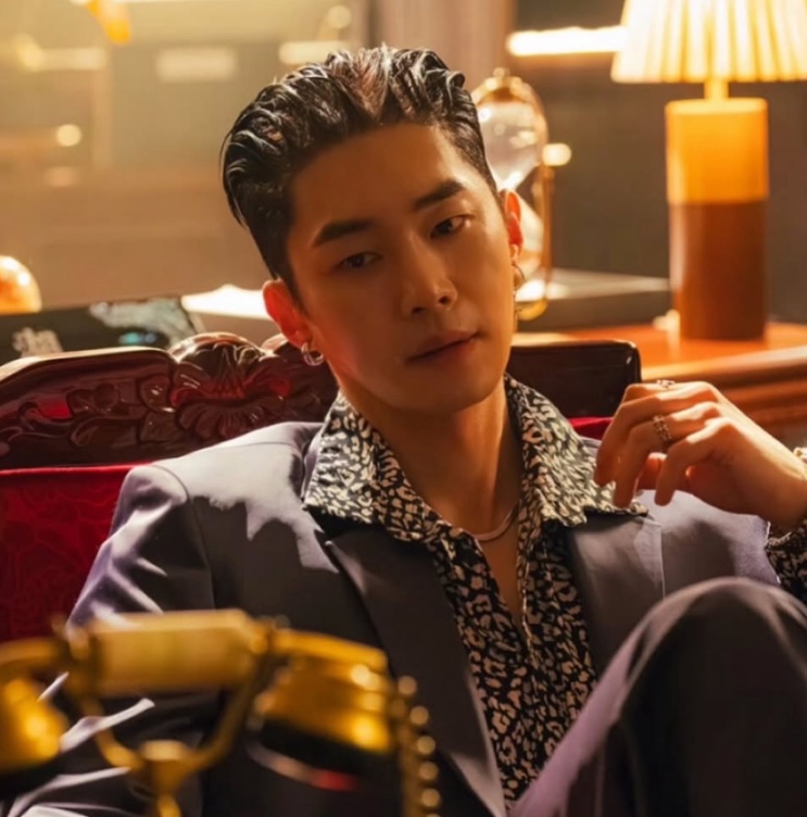 Moo Jin-sung de terno claro e camisa estampada, sentado em um sofá, como Pyo Hyeon-jun