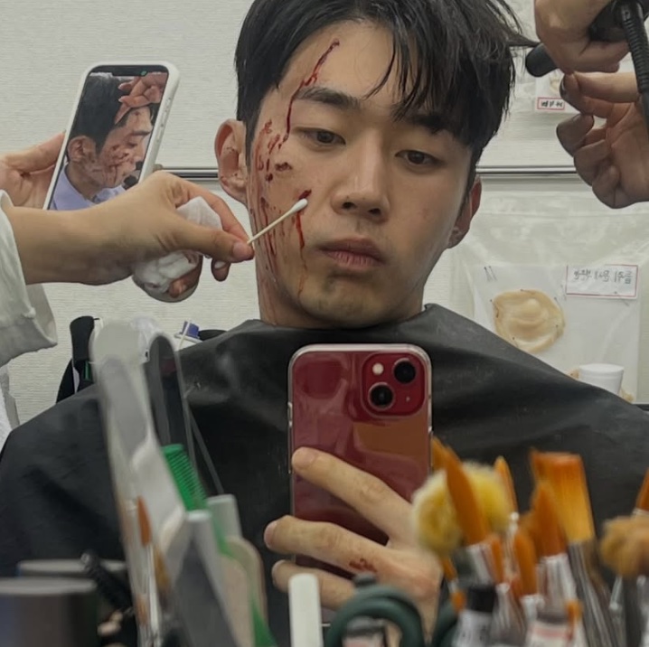 Moo Jin-sung tirando selfie no espelho com maquiagem de ferimentos no rosto, nos bastidores