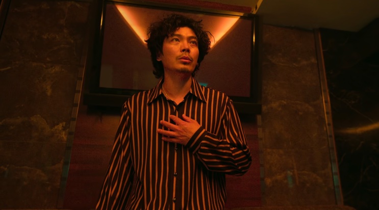 Seung-gyu de camisa listrada, com a mão no peito, em um corredor de luz alaranjada