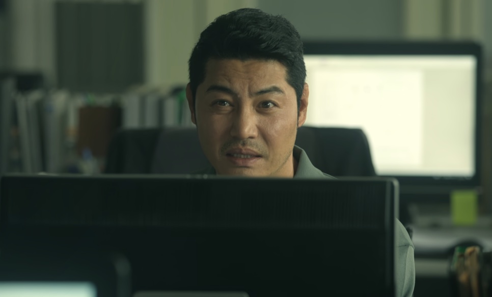 O detetive Dong-cheol sentado atrás de um computador, olhando para a frente, em O Rato