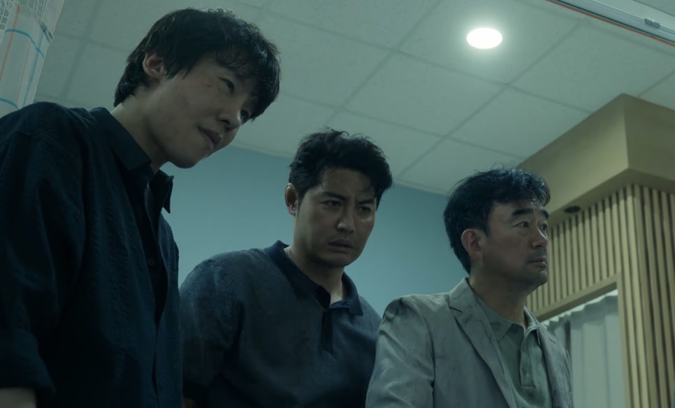 O detetive Dong-cheol entre outros dois homens, todos olhando para baixo, em um corredor claro